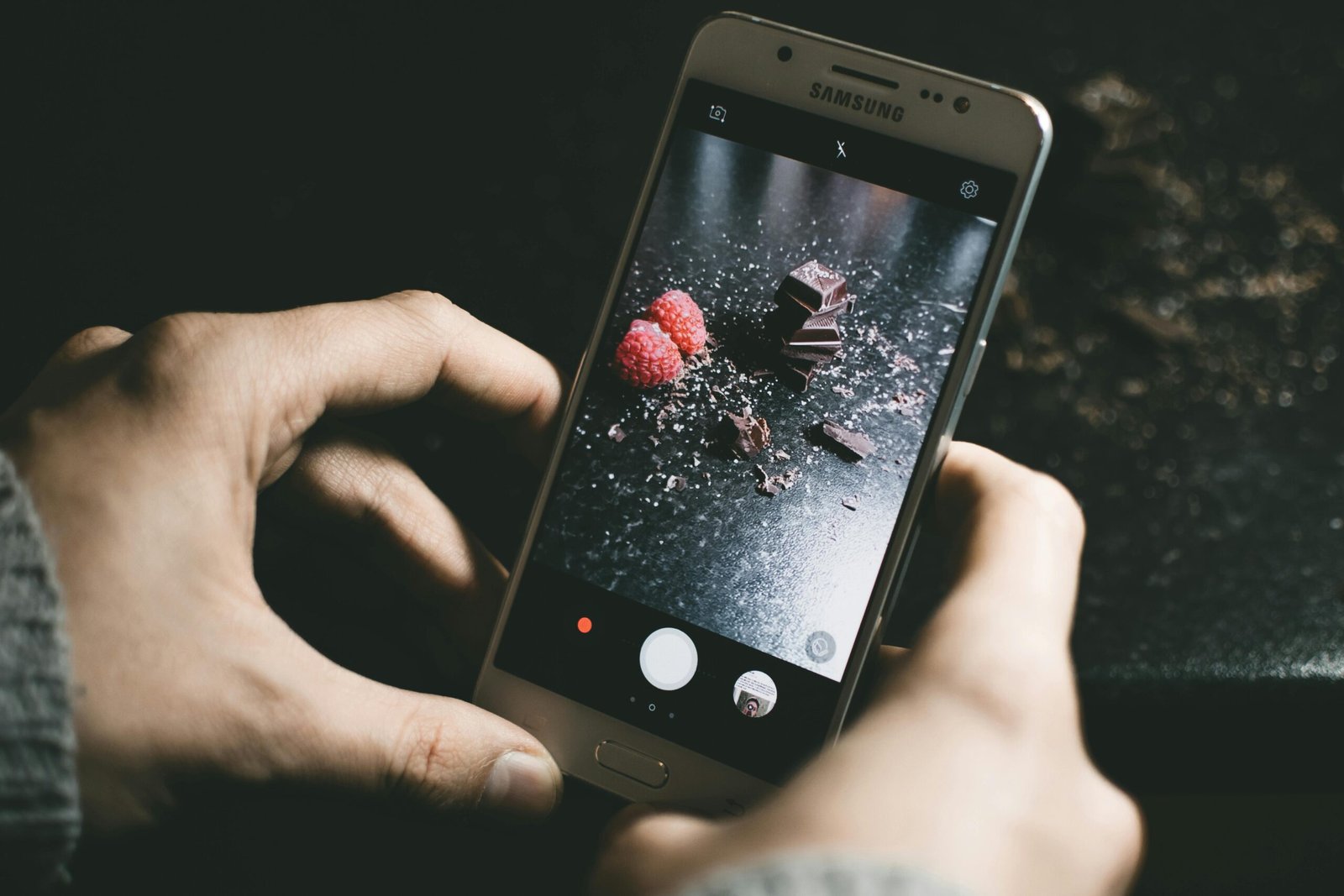 Close-up of a smartphone photographing chocolate pieces and raspberries on a dark surface.